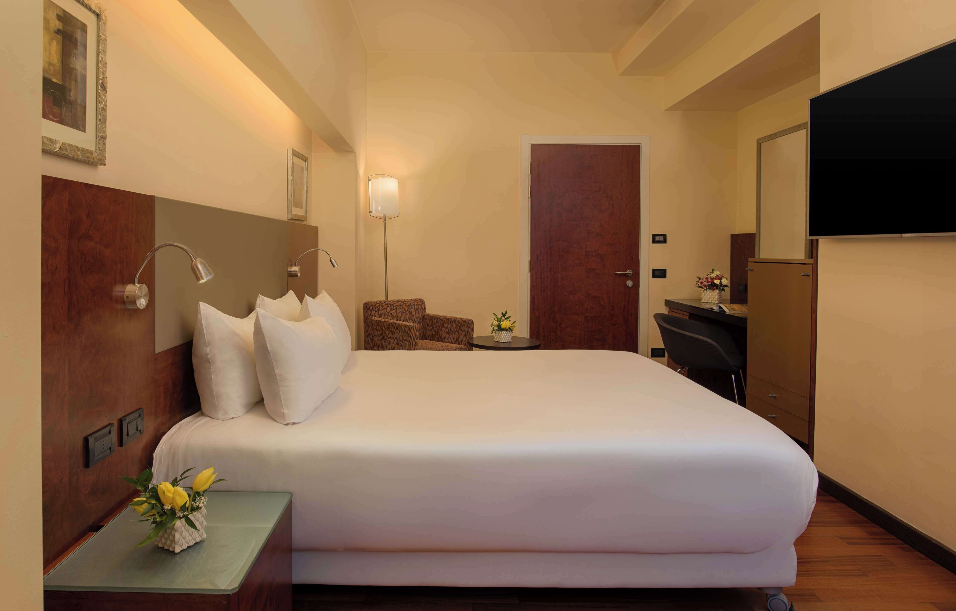 Quarto de hotel com uma cama grande, uma mesa, uma cadeira, uma mesinha com um vaso de flores, uma TV na parede e uma porta.