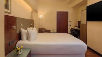 Quarto de hotel com uma cama grande, uma mesa, uma cadeira, uma mesinha com um vaso de flores, uma TV na parede e uma porta.