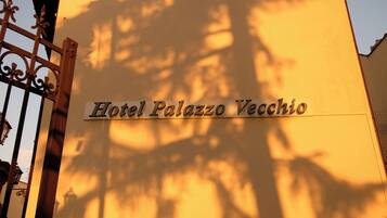 A sign for Hotel Palazzo Vecchio with a shadow of trees in the background.