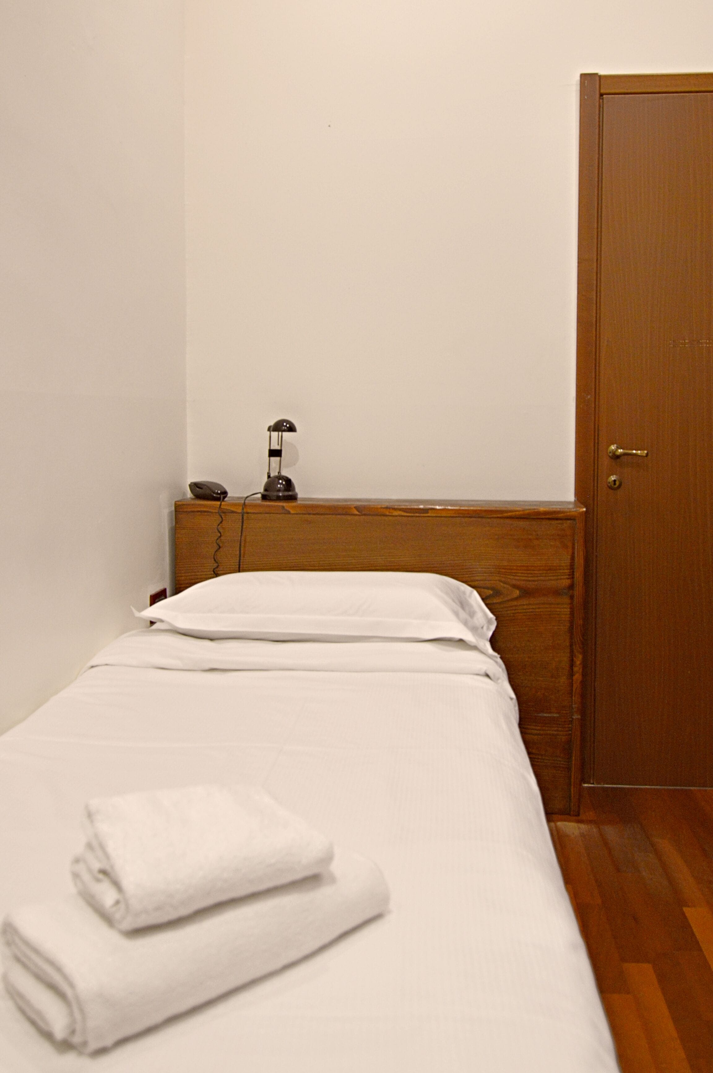 Un petit lit bien fait, avec du linge de lit blanc et une serviette pliée. Une table de chevet en bois, sur laquelle se trouvent une lampe et un téléphone, est placée à côté du lit. Une porte en bois est visible sur la droite.