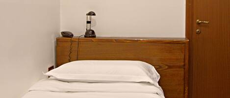 Un petit lit bien fait, avec du linge de lit blanc et une serviette pliée. Une table de chevet en bois, sur laquelle se trouvent une lampe et un téléphone, est placée à côté du lit. Une porte en bois est visible sur la droite.