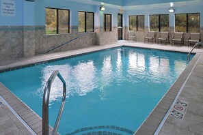 An indoor swimming pool with clear water, a lifeguard warning sign, and a seating area with chairs.
