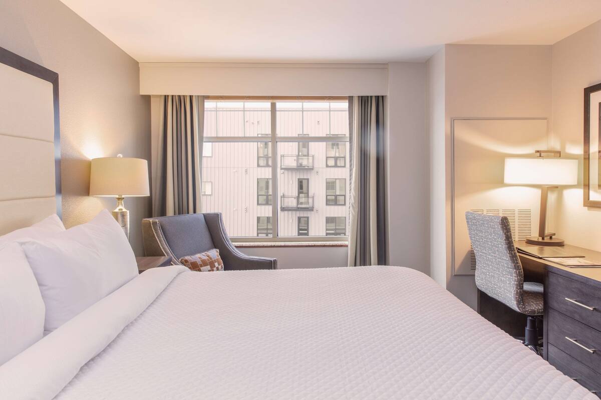 A hotel room with a large bed, a desk with a chair, a window with curtains, and a lamp on the desk.