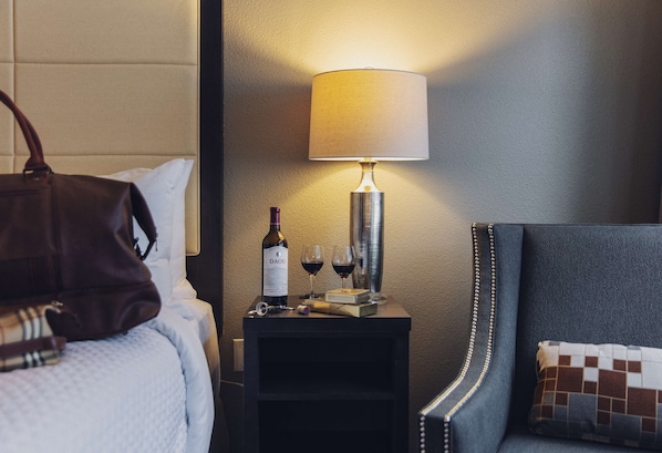 Habitación de hotel ordenada con una cama, una mesita de noche con lámpara, una botella de vino y copas.