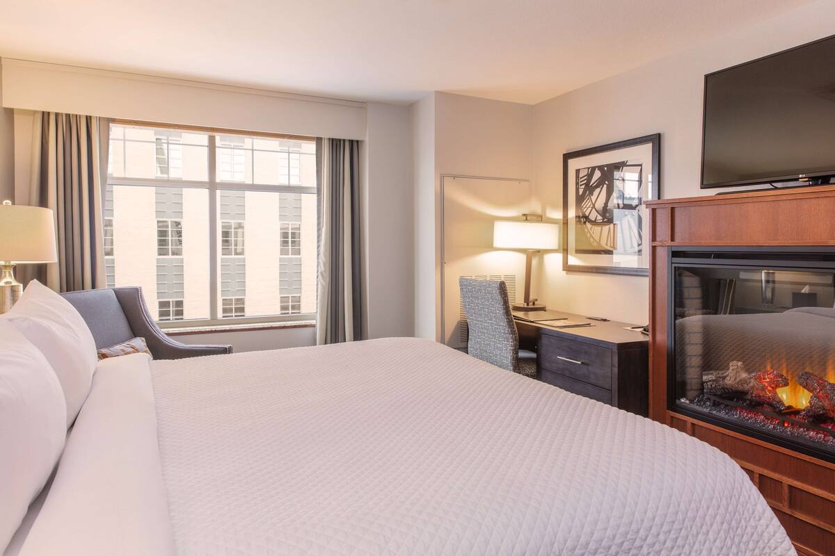 A hotel room with a large bed, a desk with a chair, a television, and a fireplace.