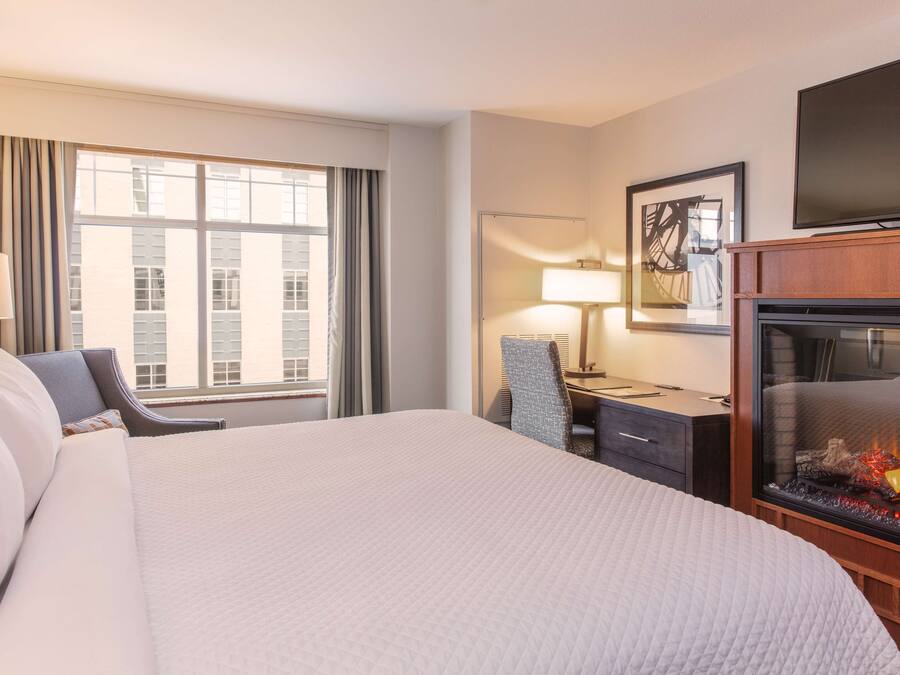 A hotel room with a large bed, a desk with a chair, a television, and a fireplace.