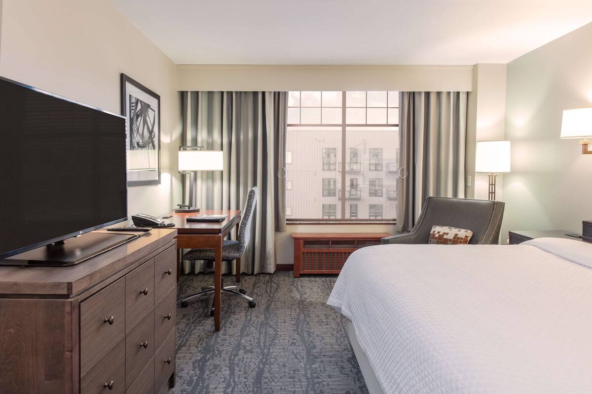 A hotel room with a bed, a desk with a chair, a television, a window with curtains, and a lamp.