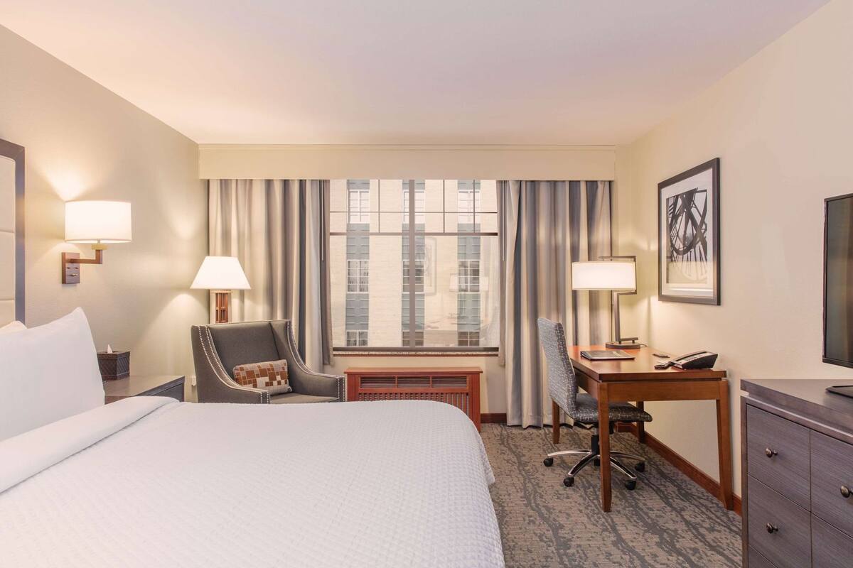 A hotel room with a bed, a desk, a chair, a desk lamp, a telephone, a picture frame, and a window with curtains.