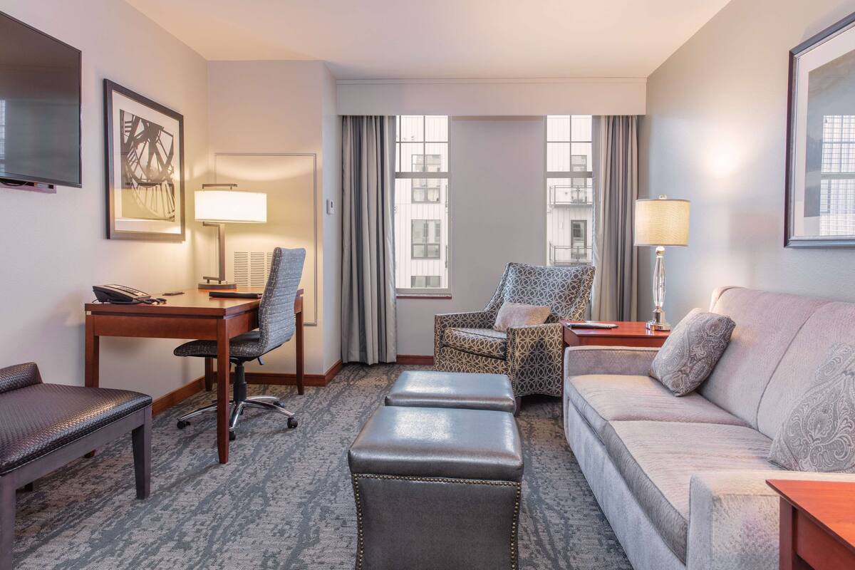A hotel room with a desk, office chair, sofa, ottoman, and a TV mounted on the wall.
