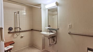 A bathroom with white tiled walls, a sink with a mirror, and a shower area with grab bars.