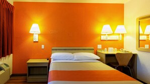 A hotel room with an orange wall, a bed with white bedding, two bedside tables with lamps, a desk with a chair, and a mirror on the wall. - Motel 6 Chico, CA (Chico)