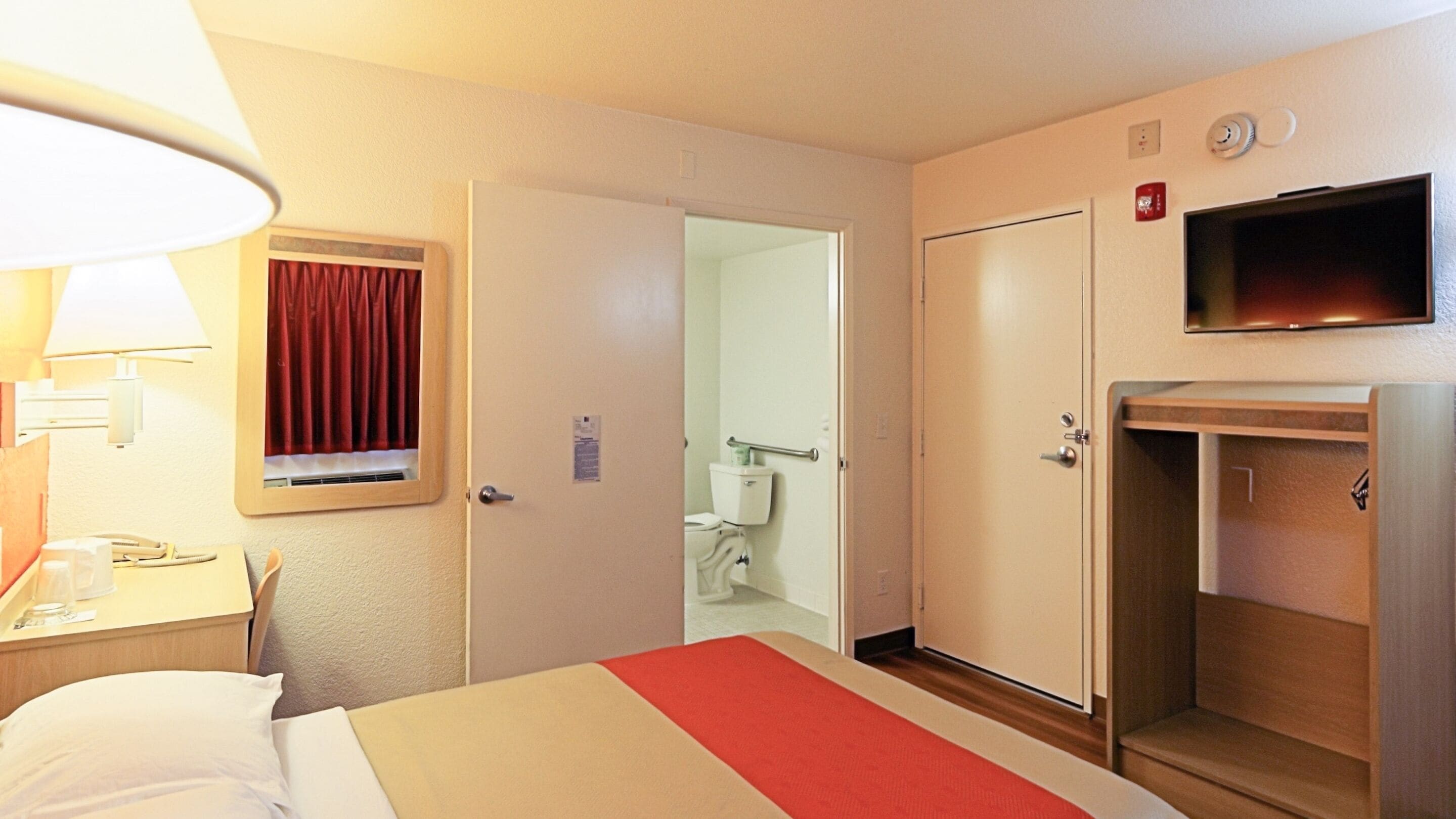 A hotel room with a bed, a desk with a lamp, a television, and a bathroom visible through an open door.