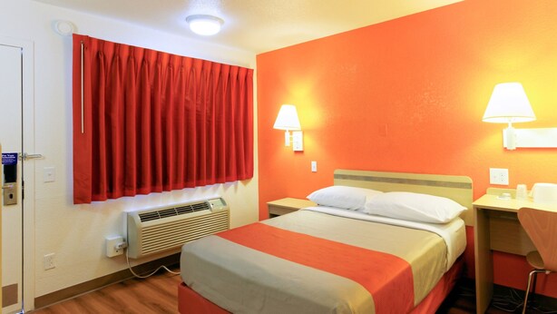 Habitación de hotel con cama, escritorio, silla, ventana con cortinas rojas, aire acondicionado y dos lámparas de pared.