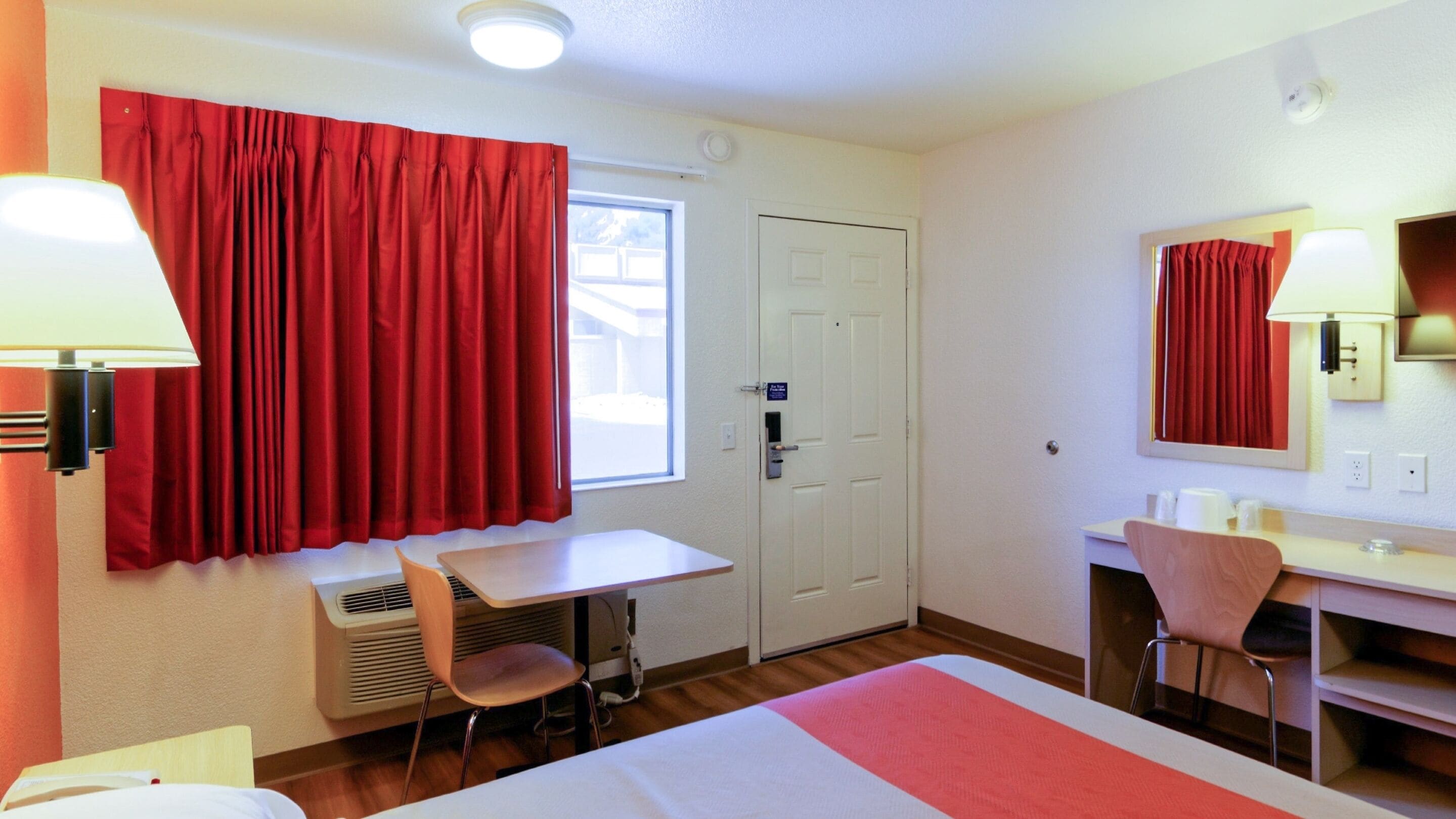 Ett hotellrum med en säng, ett skrivbord, en stol, ett fönster med röda gardiner och en dörr.
