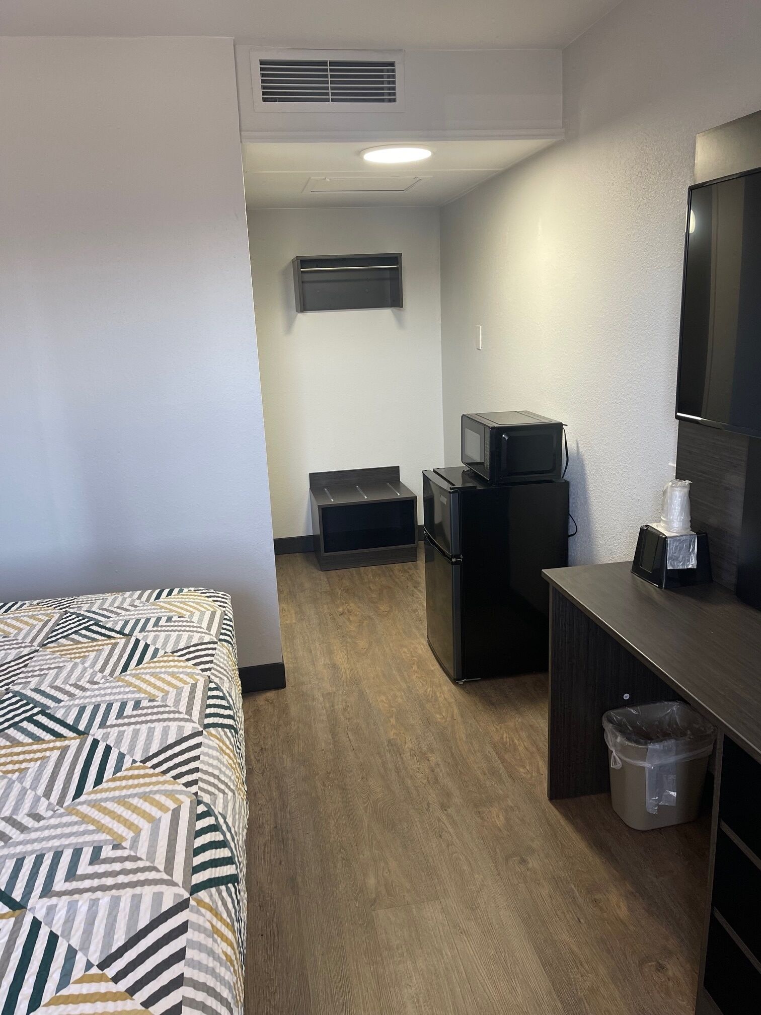 Habitación de hotel pequeña con cama, kitchenette, microondas, refrigerador, basurero y ventana.