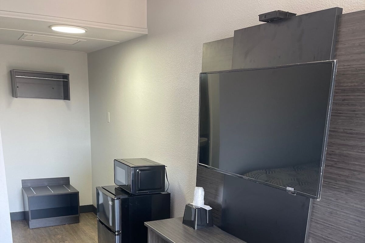A compact hotel room with a kitchenette, a flat-screen TV mounted on the wall, a small refrigerator, and a microwave.