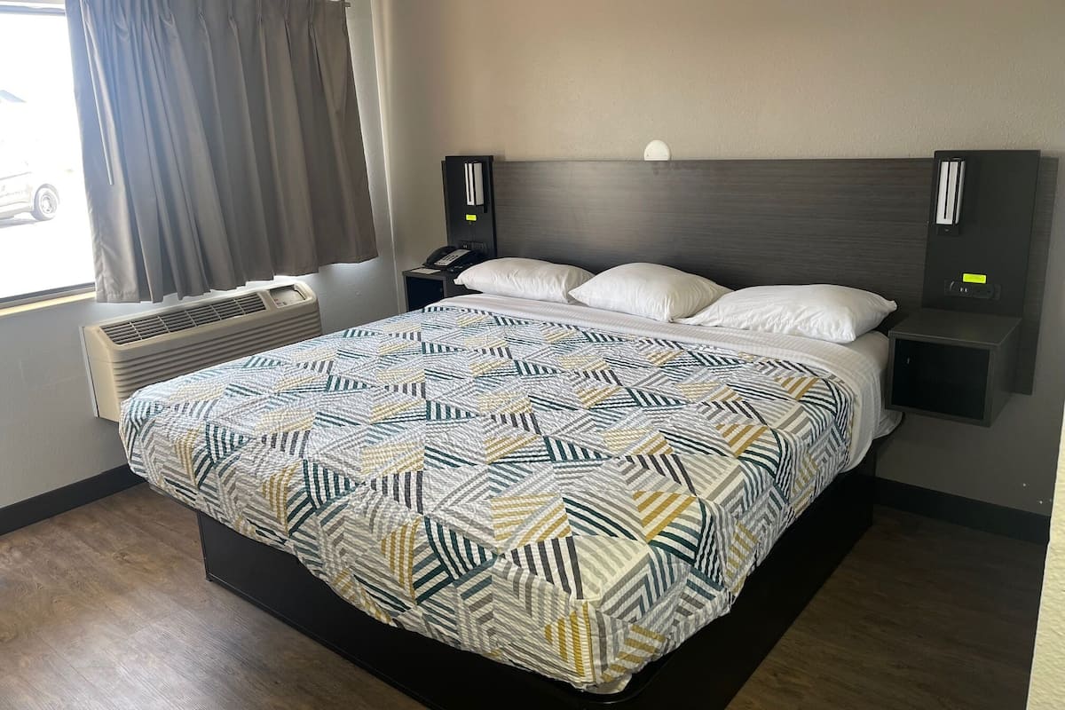 A hotel room with a large bed, a bedside table with a phone, a wall-mounted light, and a window with curtains.