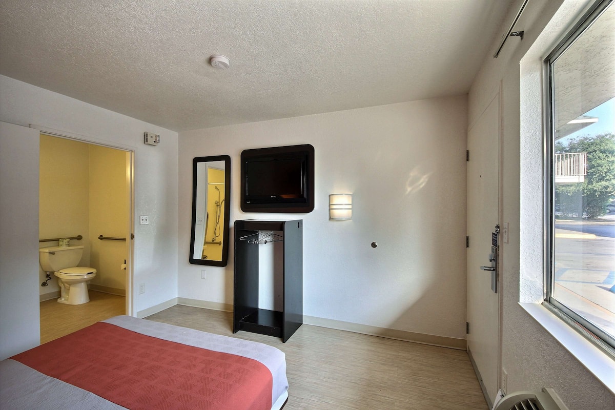 A hotel room with a bed, a TV, a mirror, a small table, and a window with a view of the outside.