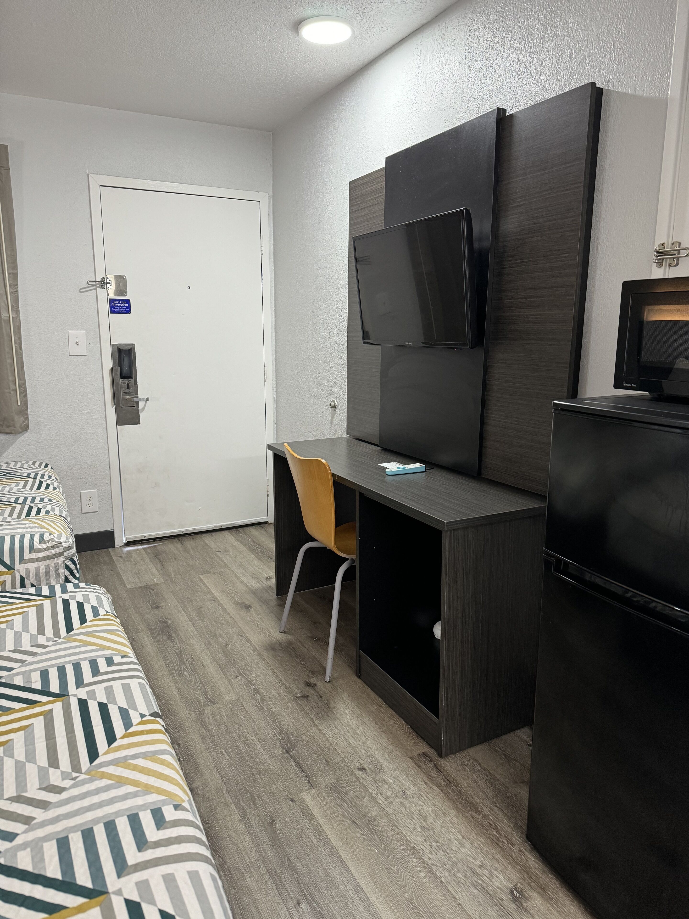 A compact hotel room with a bed, a desk, a chair, a television, and a refrigerator.