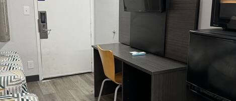 A compact hotel room with a bed, a desk, a chair, a television, and a refrigerator.