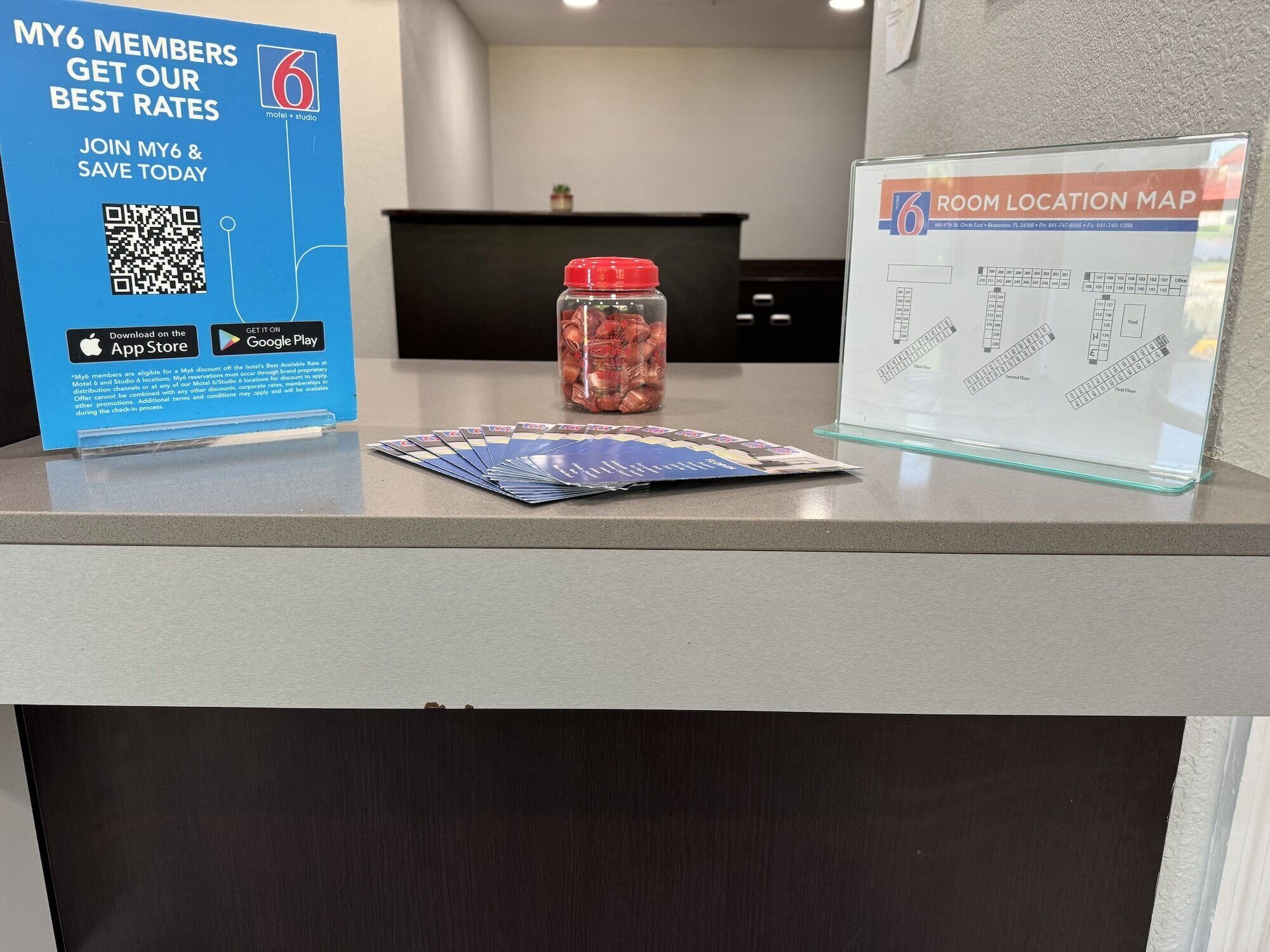 Una recepción de hotel con un cartel azul que promociona tarifas de membresía My6, un mapa de ubicación de habitaciones y un tarro de aperitivos.