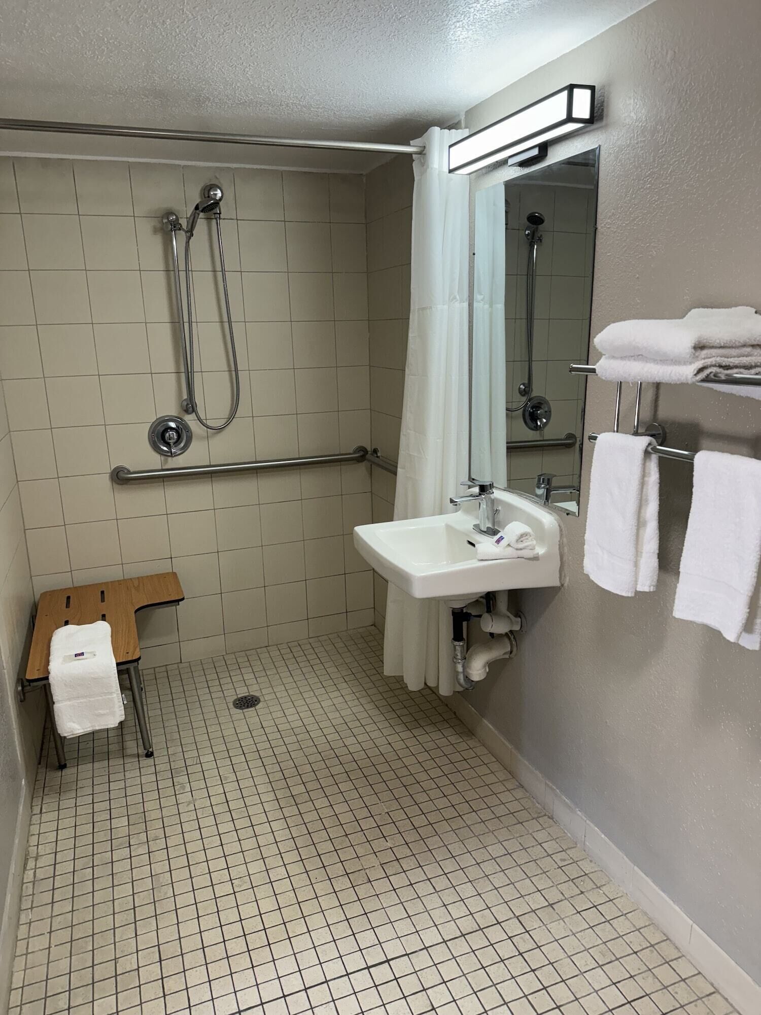 A small, tiled bathroom with a shower, a sink, and a mirror. There is a towel rack with towels and a small wooden stool with a towel.