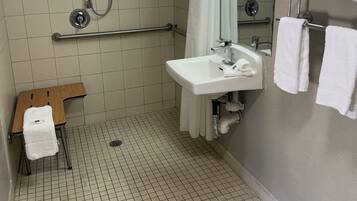 A small, tiled bathroom with a shower, a sink, and a mirror. There is a towel rack with towels and a small wooden stool with a towel.