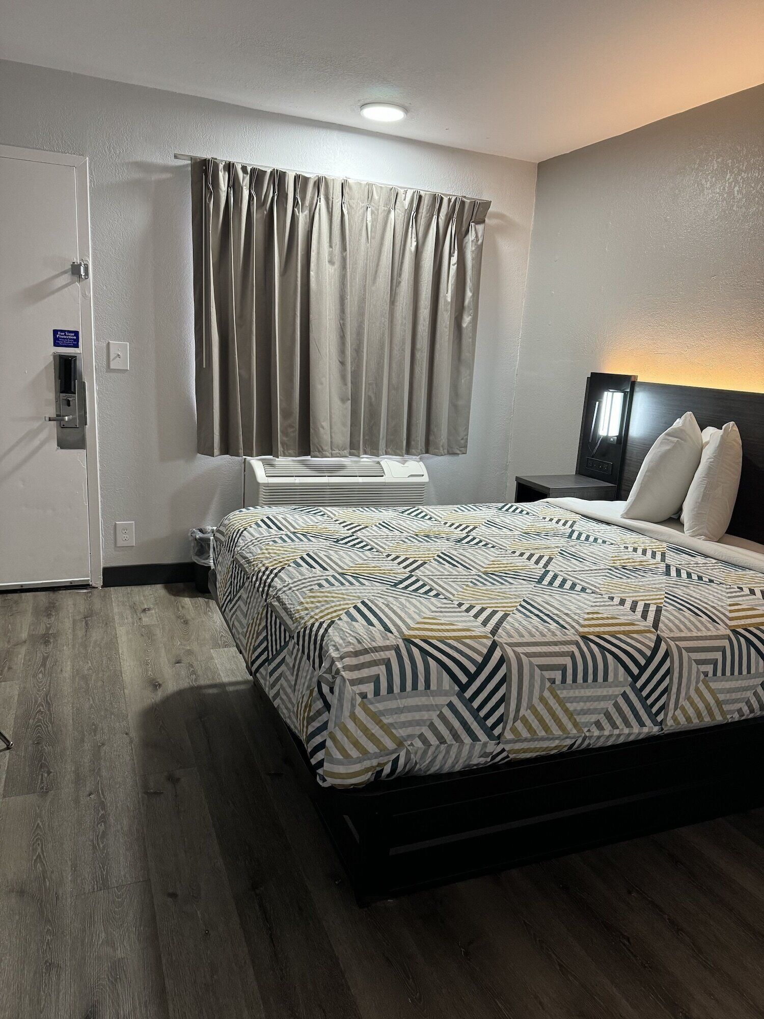 A hotel room with a bed, a nightstand, a lamp, a window with curtains, a door, and an air conditioning unit.