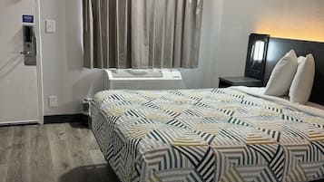 A hotel room with a bed, a nightstand, a lamp, a window with curtains, a door, and an air conditioning unit.