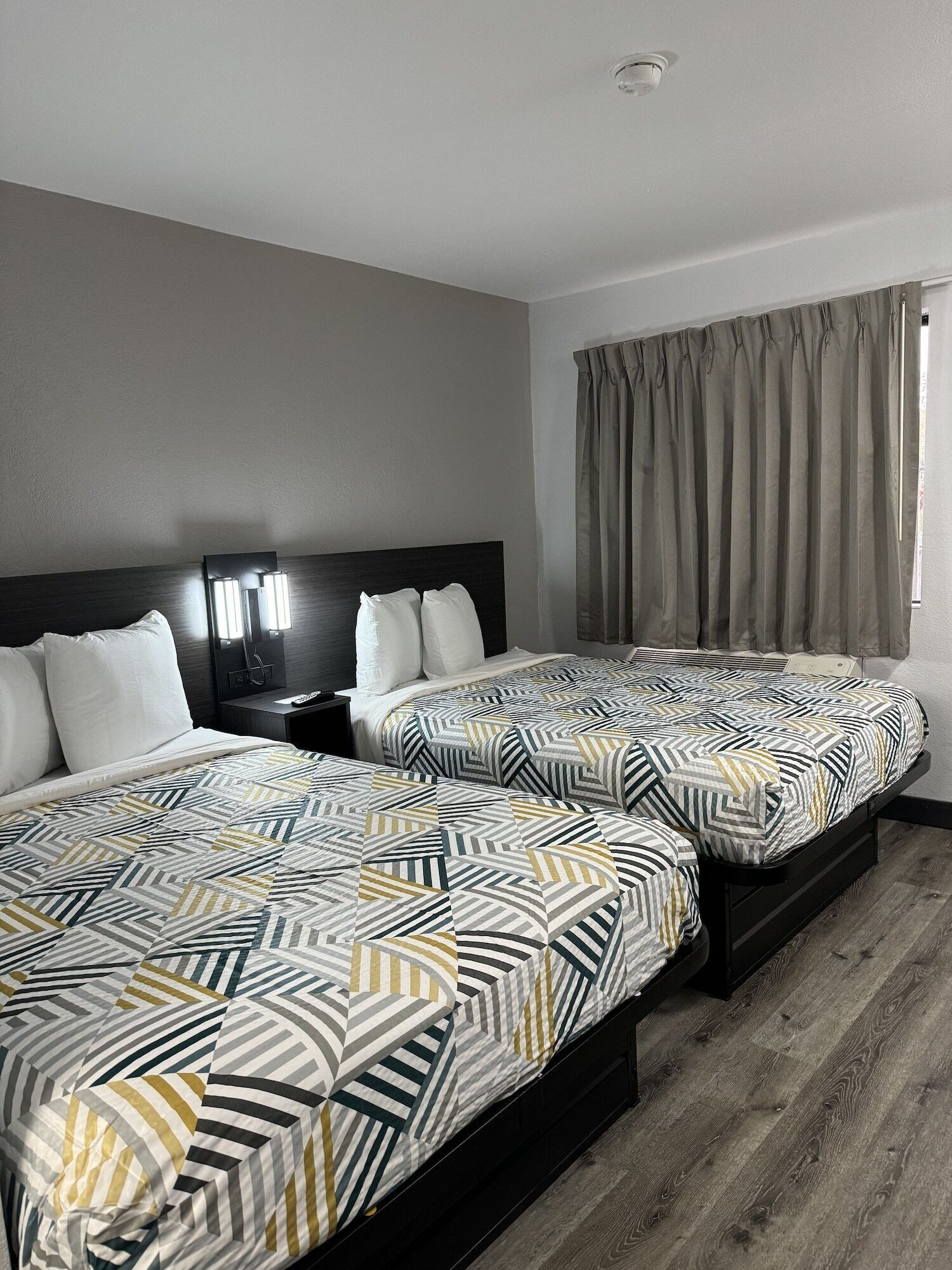 A hotel room with two beds, each with a geometric patterned bedspread, a dark headboard, and a bedside table with a lamp.