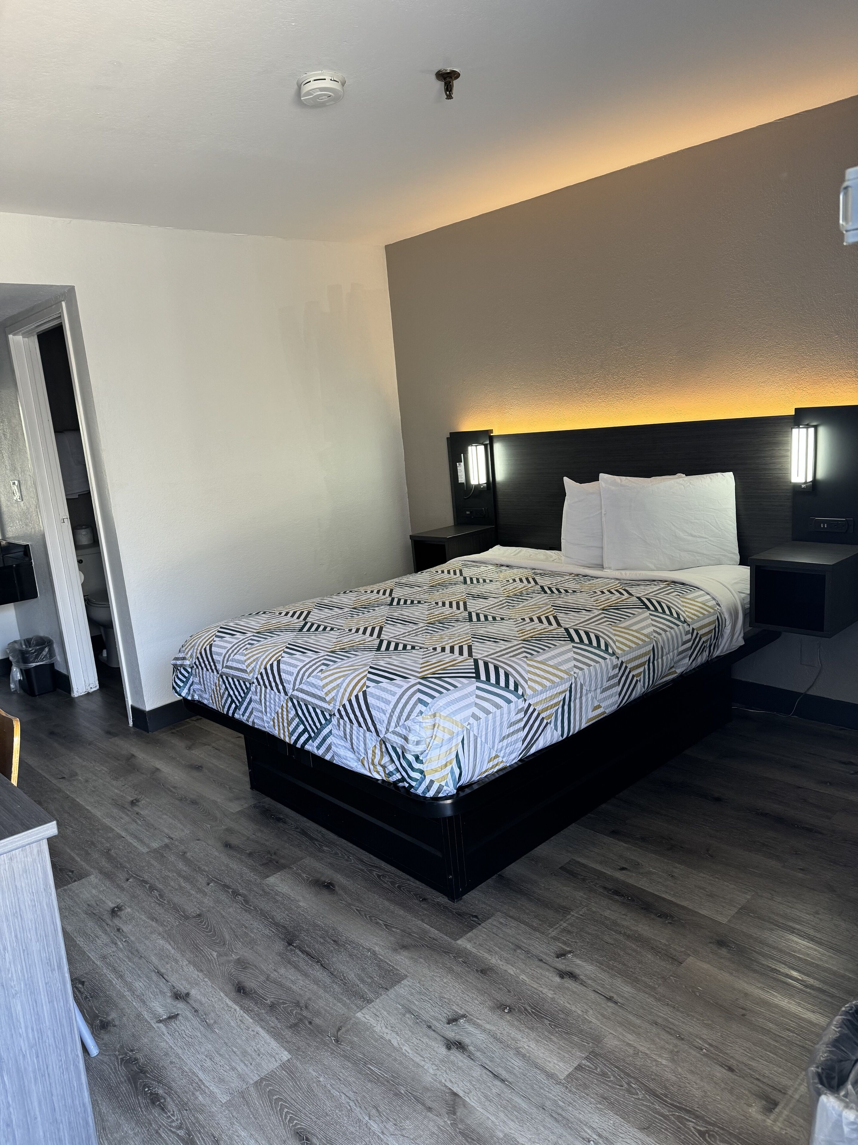 A modern hotel room with a large bed, a bedside table, a nightstand, a wall-mounted light, and a ceiling light.