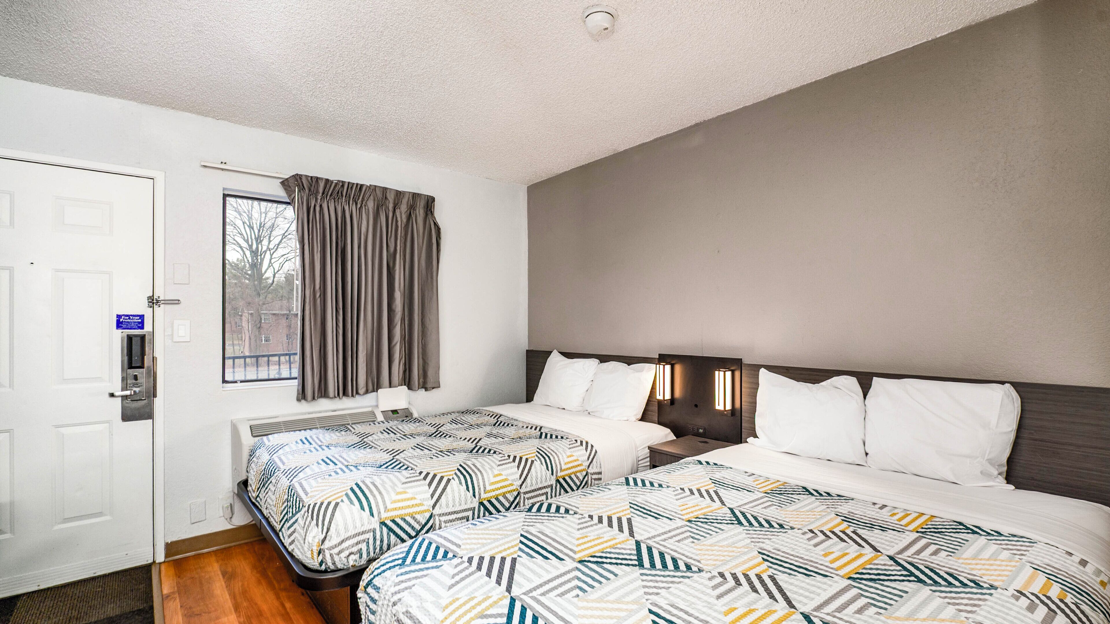 A hotel room with two beds, a window with curtains, a door with a keycard reader, and a thermostat on the wall.