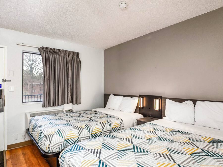 A hotel room with two beds, a window with curtains, a door with a keycard reader, and a thermostat on the wall.