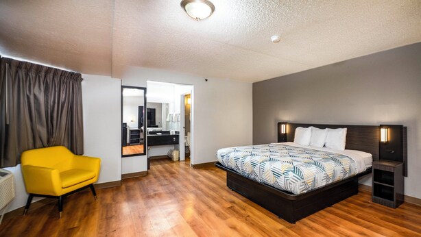 A hotel room with a large bed, a yellow armchair, a desk with a chair, and a bathroom visible through an open door. - Motel 6 Wethersfield, CT - Hartford (Wethersfield)