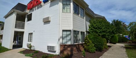 Un motel à deux étages, avec une enseigne « Motel 6 », un balcon au deuxième étage et un jardin bien entretenu avec des arbustes et un trottoir.