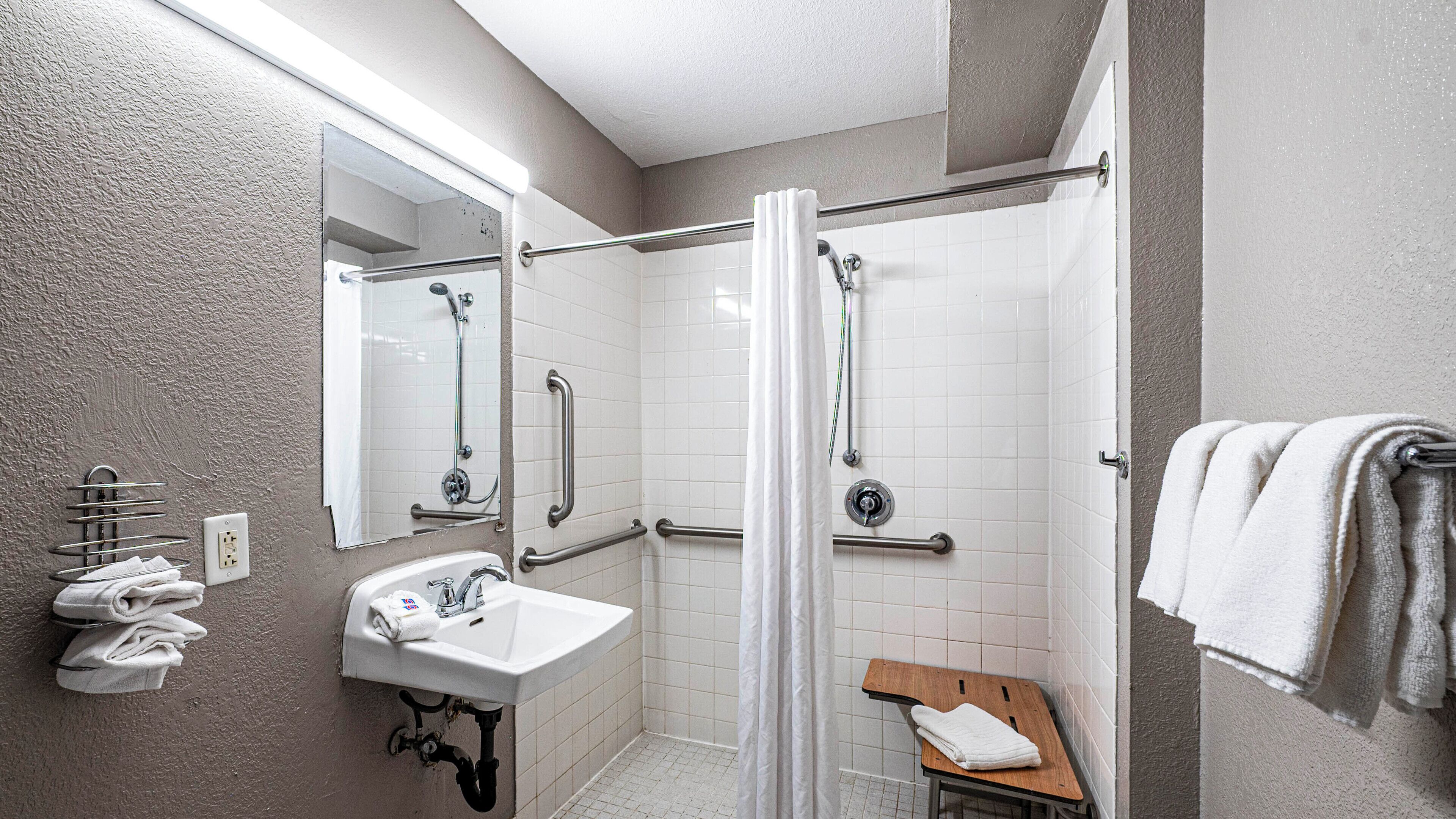 A bathroom with a sink, mirror, shower, and handrails.