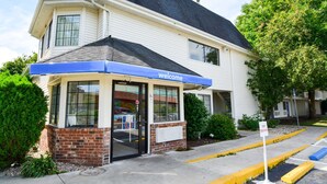 A hotel entrance with a 'Welcome' sign, a brick facade, and a parking area with yellow curbs. - Motel 6 Wethersfield, CT - Hartford (Wethersfield)