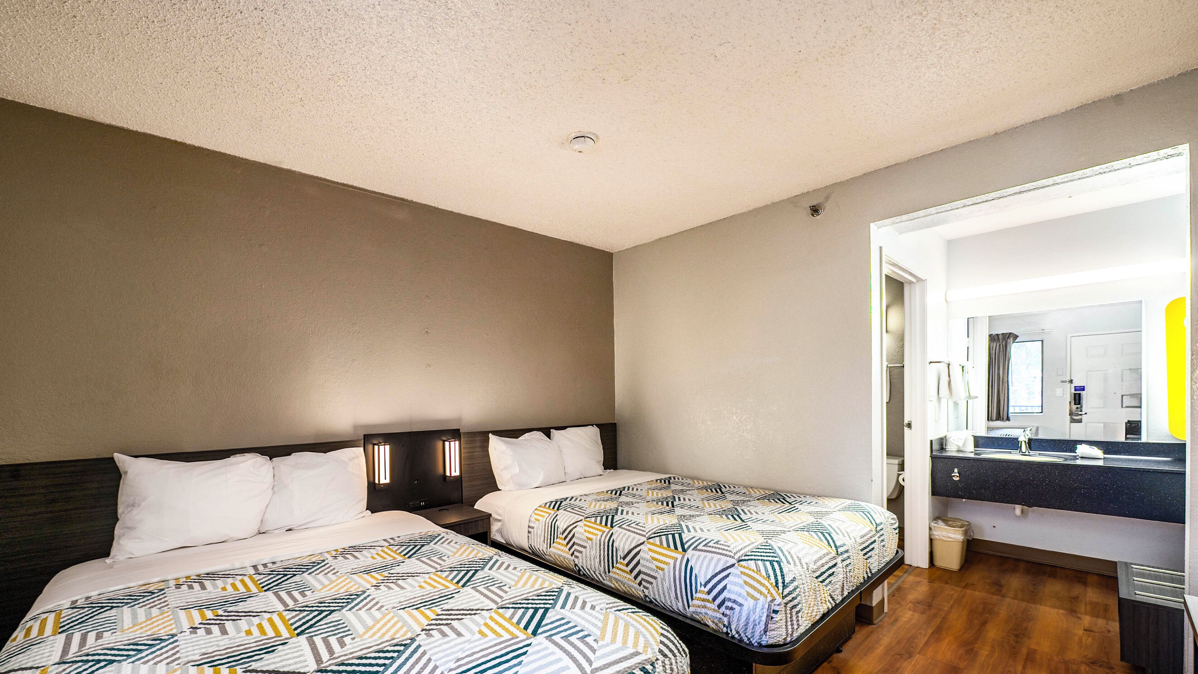 A hotel room with two beds, a bathroom with a mirror and sink, and a wooden floor.