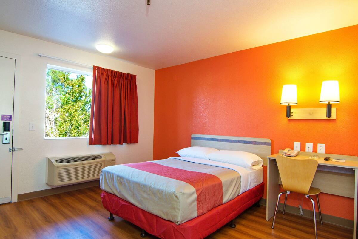 A hotel room with a bed, a desk with a chair, a window with curtains, and a wall-mounted lamp.
