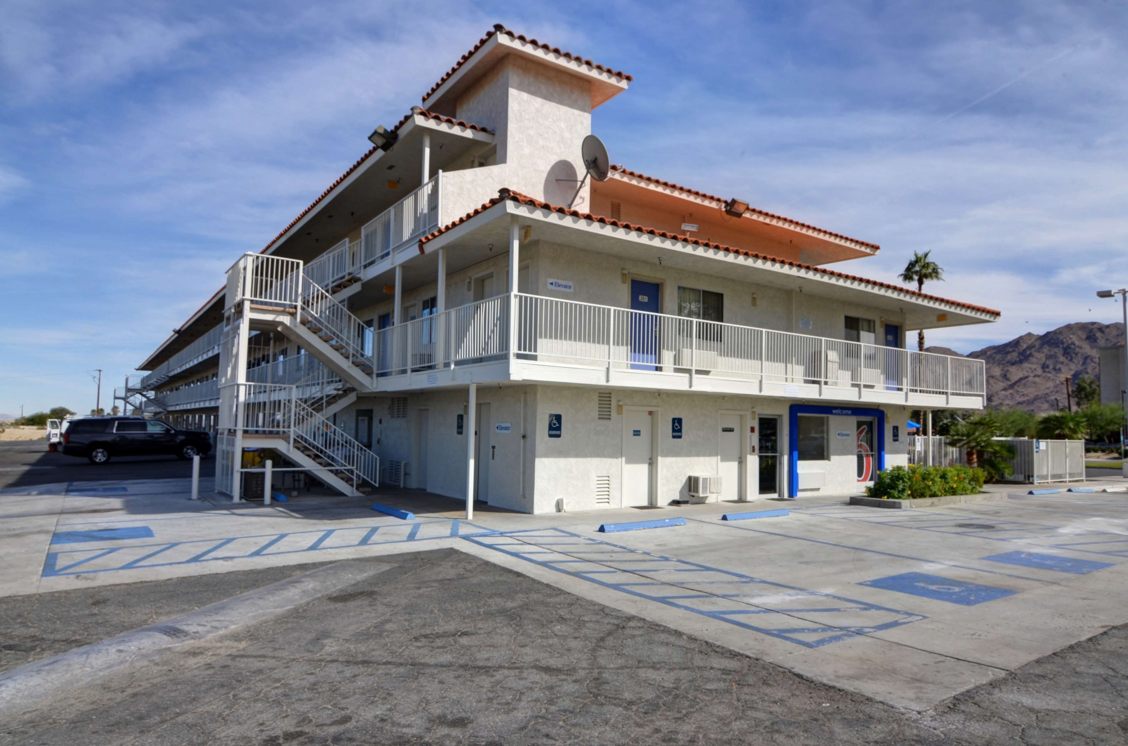 Un edificio de varios pisos con techo de tejas rojas, una rampa accesible para sillas de ruedas y un estacionamiento con señalización azul para discapacitados.
