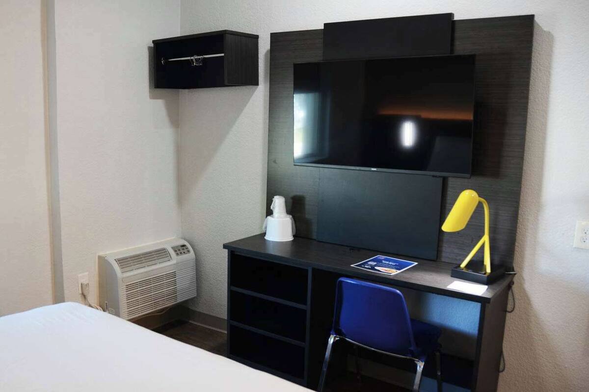 A hotel room with a bed, a desk with a chair, a TV, a heater, and a small shelf with a lamp.
