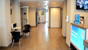 A hotel corridor with a reception desk, a microwave, two chairs, and a TV screen displaying a promotional message. - Motel 6 West Des Moines, IA (West Des Moines)