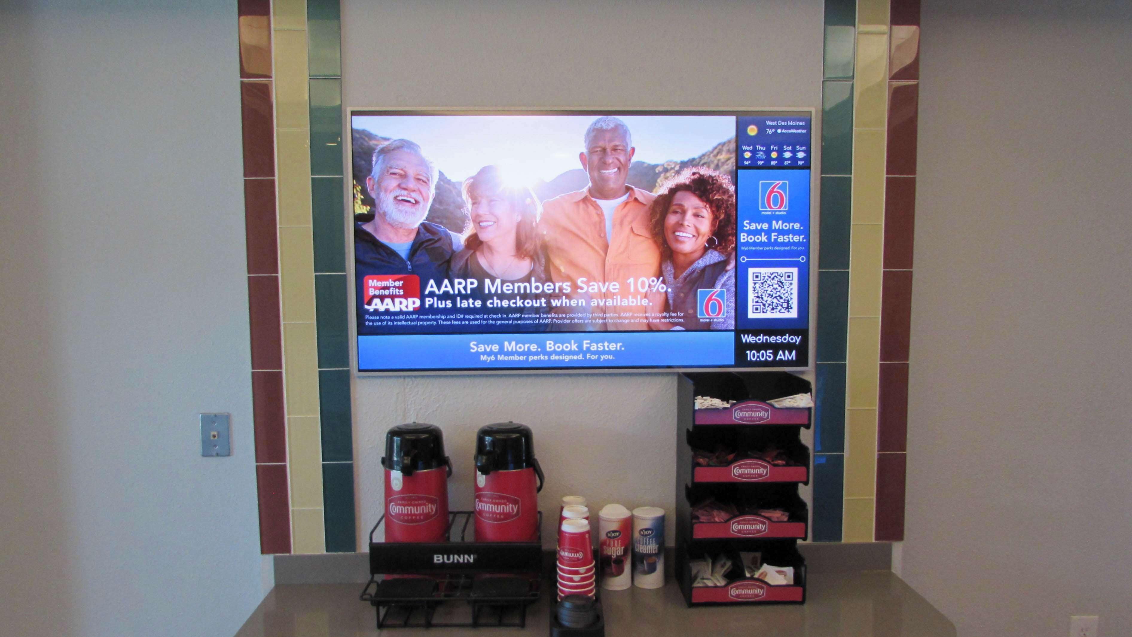 Un téléviseur diffusant un message publicitaire destiné aux membres d’AARP, avec un coin café en dessous, comprenant des tasses à café et une machine à café portant le logo de l’entreprise.