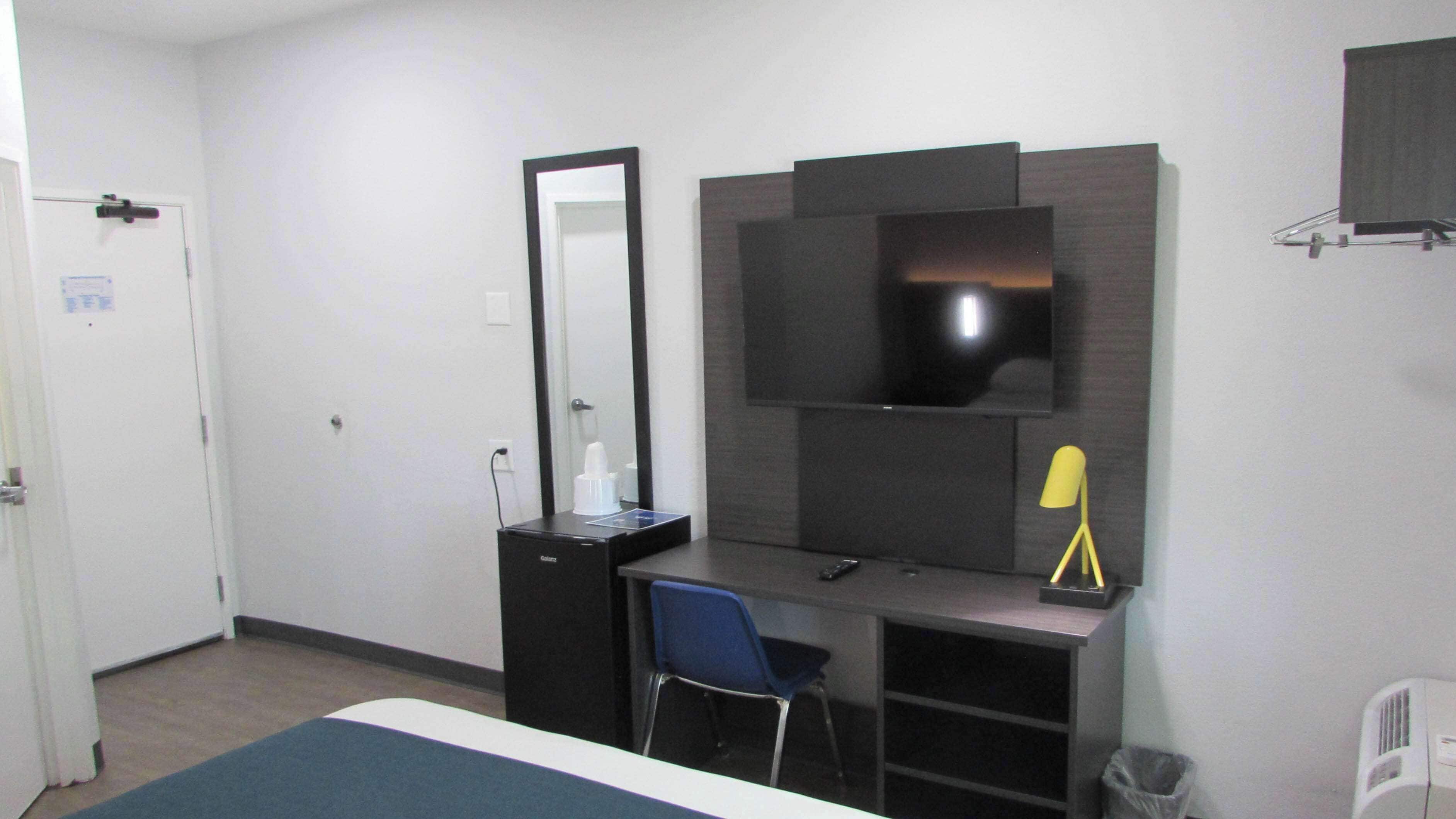 A hotel room with a bed, a desk with a chair, a mirror, a TV, and a trash bin.