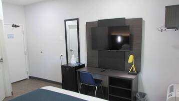 A hotel room with a bed, a desk with a chair, a mirror, a TV, and a trash bin.
