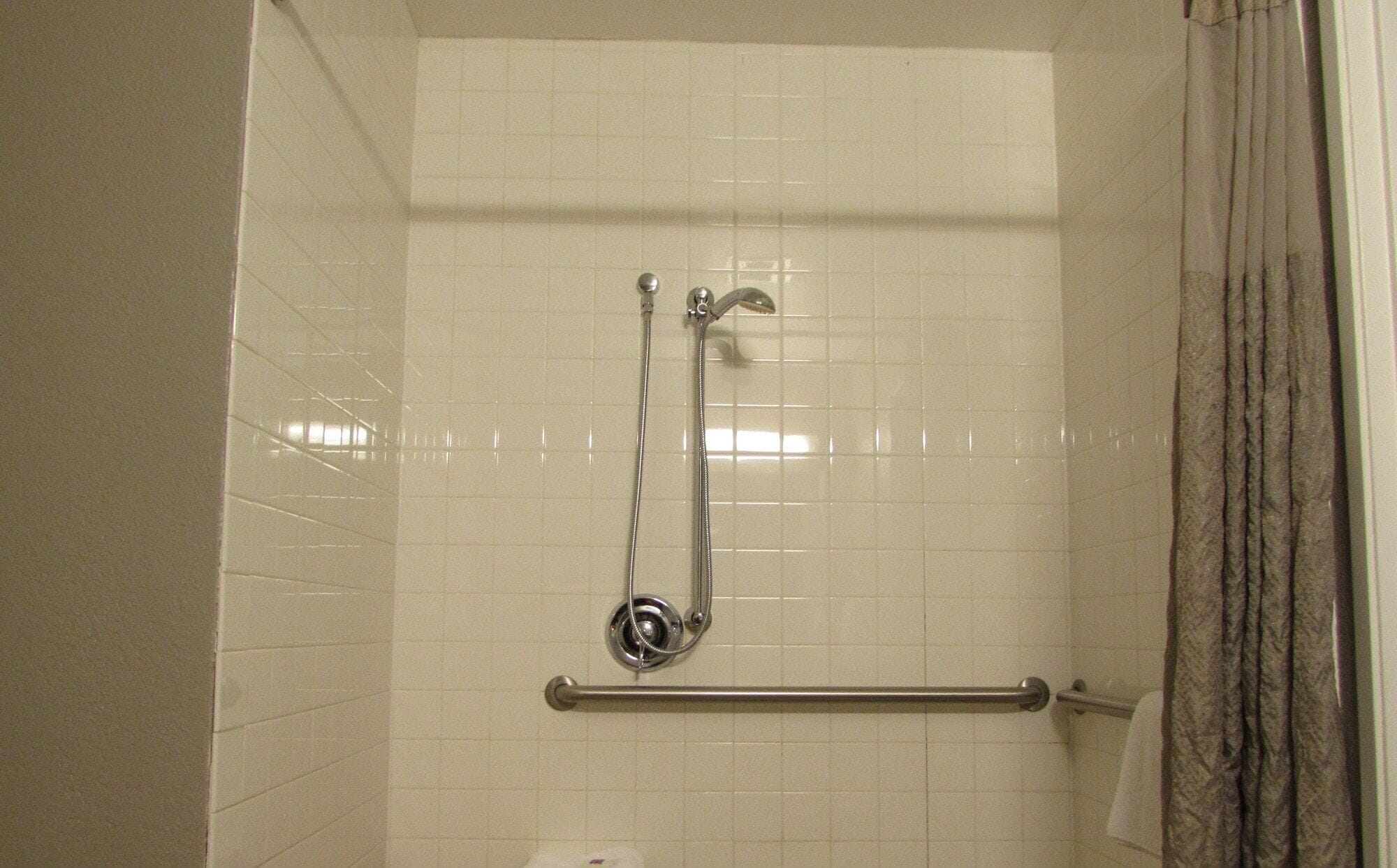 Een douche met een handgreep, witte betegelde wanden en een gordijn.