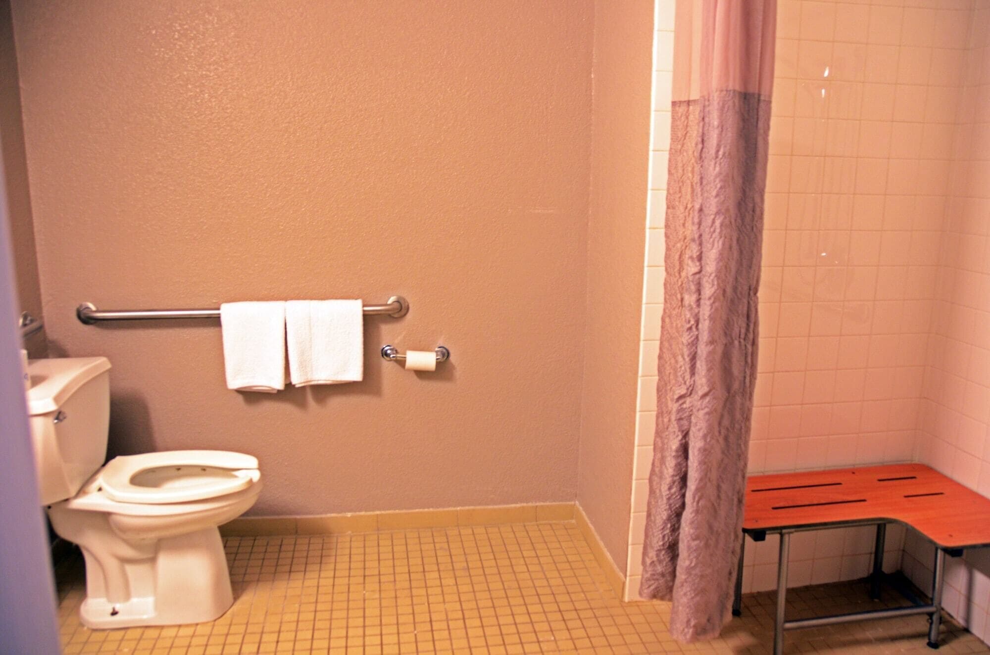 A small bathroom with a toilet, a towel rack with two towels, a shower curtain, and a bench.