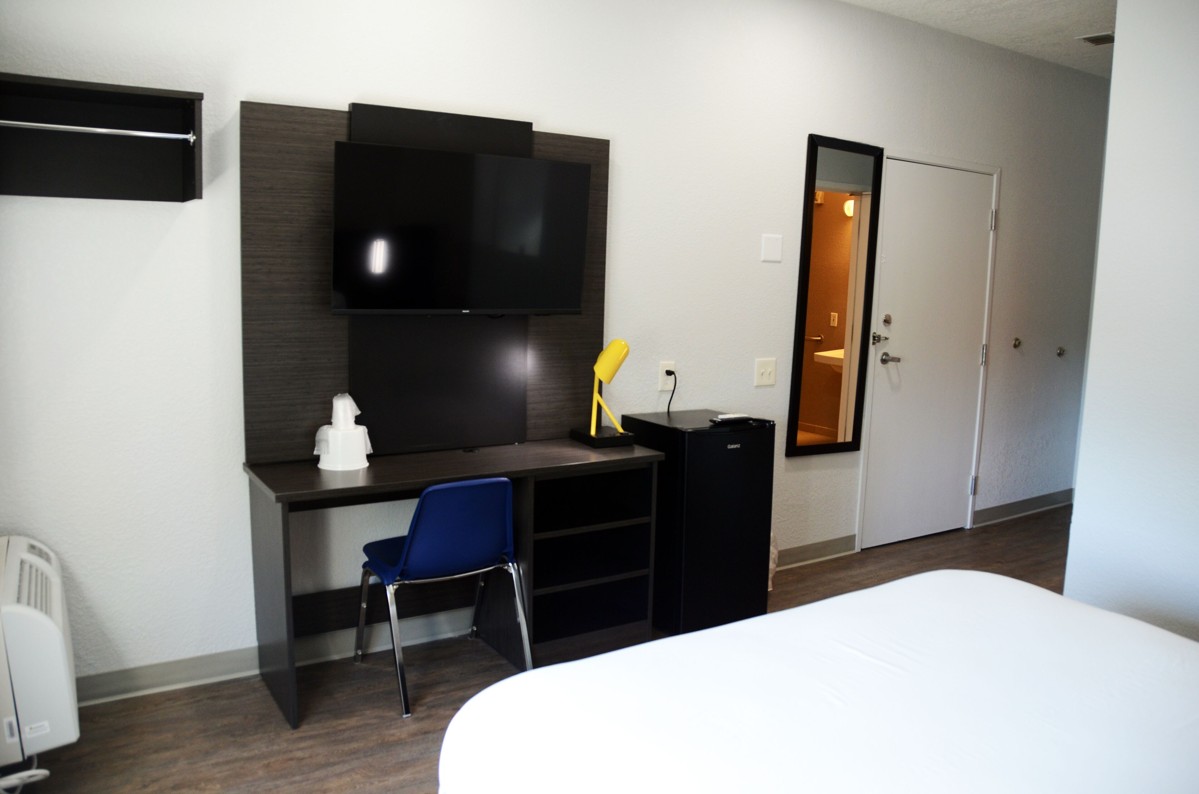 A hotel room with a bed, a desk with a chair, a TV mounted on the wall, a small black cabinet, and a door leading to a bathroom.