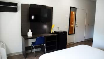 A hotel room with a bed, a desk with a chair, a TV mounted on the wall, a small black cabinet, and a door leading to a bathroom.