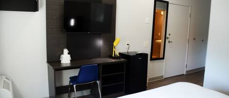 Habitación de hotel con una cama, un escritorio con silla, un televisor montado en la pared, un mueble negro pequeño y una puerta que da al baño.